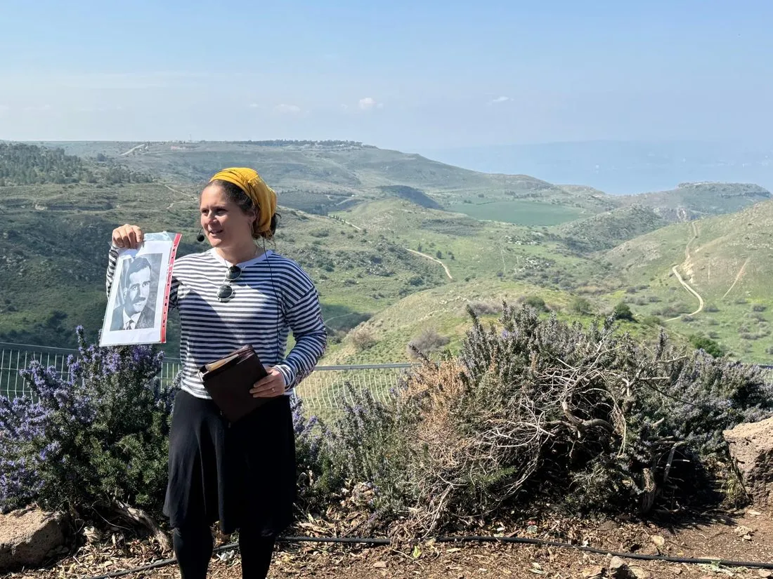 Orit Kropp guiding a tour outdoors, holding up a photograph, with green hills and a lake behind her